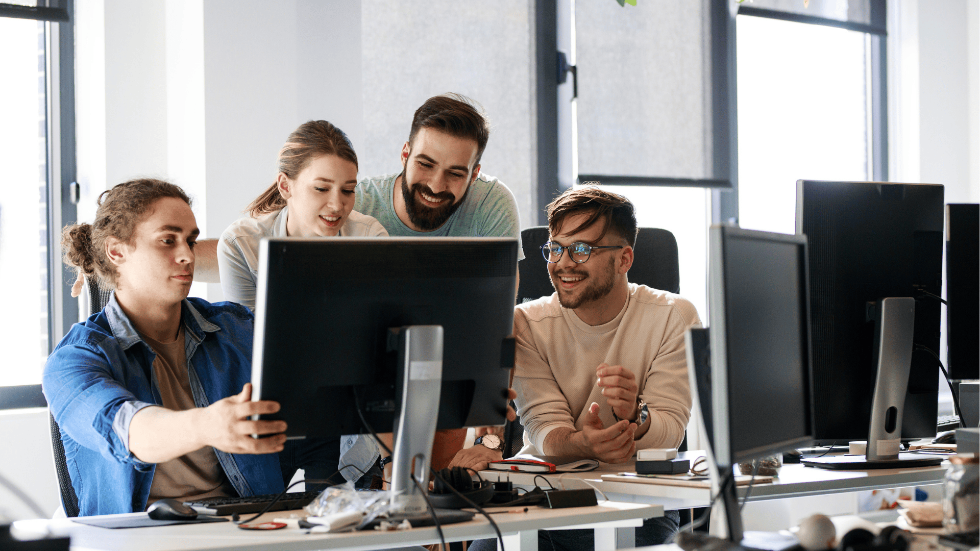 Hier werden vier Personen im Bürokontext gezeigt. Sie arbeiten zusammen und schauen auf einen Monitor. Es wird ein beispielhafter Arbeitsplatz für Entwickler*innen dargestellt.